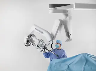 A medical professional in scrubs and a mask operates a large robotic surgical arm above a patient covered with a blue drape in a bright, sterile environment.