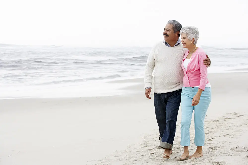 An older couple walks barefoot on the beach, smiling and holding each other. The man wears a white sweater and jeans, and the woman wears a pink cardigan and light blue pants. The ocean waves are in the background.